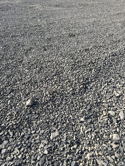 The image displays a close-up, ground-level perspective of a surface covered in loose, gray gravel. The gravel pieces are of varying sizes and shapes, appearing sharp and angular. The texture is rough and uneven. The lighting suggests an outdoor environment, likely during daylight hours, with shadows cast by the individual stones. The scene is devoid of any people or specific landmarks, presenting a simple, textural study of the gravel. There is no information within the image to indicate the location of Izyayu, Russia.