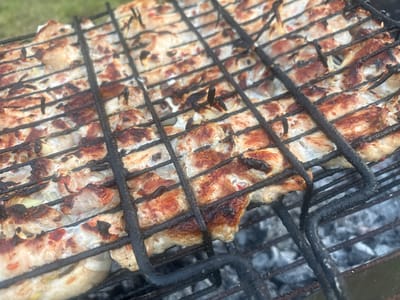 The image shows a close-up of food being grilled on a wire rack over glowing coals. The food appears to be pieces of meat, possibly chicken or pork, with some darker, charred areas indicating it is being cooked. The meat is arranged in rows across the grill. The background is slightly blurred, showing glimpses of green grass and gray ash from the coals. The lighting suggests it is daytime, possibly outdoors in a natural setting. No people are visible in the frame, and there is no visible text. The scene depicts the activity of barbecuing or grilling, a common outdoor cooking method. The context provided, "Цярэшкавіцкі сельскі Савет, Belarus," suggests the location of this activity.