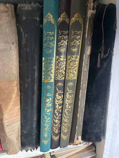 The image shows a close-up, top-down view of several old books standing upright on a shelf. The books are arranged from left to right: a thick, beige-colored book with a rough texture; a black book with a worn leather binding; a teal book with gold Arabic calligraphy and floral designs; a dark purple book with similar gold Arabic calligraphy and designs; and a gray book with gold Arabic calligraphy and designs. The books appear to be old and well-used, with some signs of wear and tear on their bindings. The shelf itself is not fully visible, but appears to be white and slightly textured. Below the upright books, other books or papers are visible, stacked horizontally. The scene is indoors, with soft lighting. There are no people or animals present. The location is not specified in the image or prompt, but the prompt asks to include "the location in the city of Ташлак, Kyrgyzstan." However, there are no visual cues in the image that indicate this specific location.