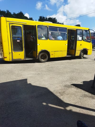 A yellow, aging bus with open doors is parked on a paved surface, likely a street or parking lot, under a partly cloudy sky. The bus has several passengers visible through the windows. Some people are seated inside, and one person appears to be standing near the open front door. The bus shows signs of wear, with rust visible on the bodywork, particularly around the wheel arches and door frames. The ground is asphalt, with a distinct shadow cast by the bus and other unseen objects. The overall scene suggests a typical day in a town or city, with the bus likely serving as local public transportation. The location is identified as Berestyn, Ukraine.
