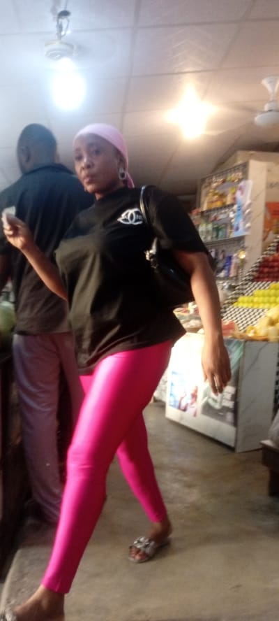 A woman wearing a black t-shirt with a white logo, bright pink leggings, and silver sandals is standing in what appears to be a small retail establishment. She also has a pink head covering. There's a man standing beside her to her left. The background features shelves stocked with merchandise and what looks like a display of fruit. There are also fluorescent lights hanging from the ceiling. The location is identified as Rijiyar Zaki, Nigeria.