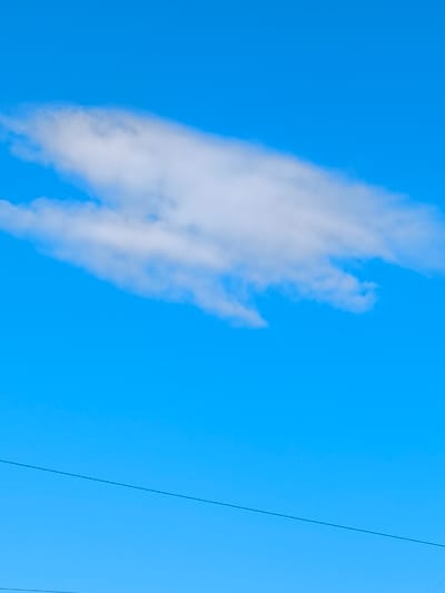 A clear, bright blue sky dominates the image, with a single, wispy white cloud stretching horizontally across the upper third. Below the cloud, a thin, dark horizontal line, likely a power line, bisects the lower portion of the frame. The scene is characteristic of an outdoor environment, possibly in the city of Abay, Kazakhstan, during daylight hours. No people or specific landmarks are visible.