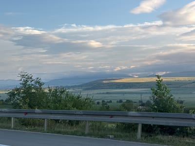 This is a landscape photograph taken from a roadside, looking out over rolling hills and fields under a cloudy sky. In the foreground, a grey metal guardrail runs horizontally across the image, with its metal posts visible. Beyond the guardrail, lush green trees and bushes are prominent. The middle ground features vast agricultural fields, characterized by distinct rows and patterns, stretching towards a gently sloping hill. The hill is illuminated by sunlight breaking through the clouds, casting a golden glow on its upper portion, while the lower parts are in shadow. In the distance, hazy mountains are visible against the sky. The sky itself is a mix of blue and white clouds, with some areas showing brighter, sunlit patches. The overall impression is a rural, natural setting, likely in the vicinity of Akhalsheni, Georgia, given the context. There are no people or animals visible in the image.