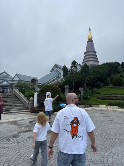 In Tambon Ban Luang, Thailand, a man, a child, and another man are walking on a paved circular courtyard. The man in the foreground is facing away from the camera, wearing a white t-shirt with an embroidered orange scooter and the words "I'm On My Way" on the back. He is also wearing blue jeans and a watch on his left wrist. To his left, a child with blonde hair is walking ahead, also facing away from the camera. The child is wearing a white t-shirt and grey pants with a smiley face pattern. Behind the child, another man in a white t-shirt and blue shorts is walking away from the camera, pointing to his right.

In the background, a large, multi-tiered pagoda with a golden spire stands prominently against an overcast sky. Several buildings with gray roofs are visible on the left, and a lush, green hillside with manicured flower beds is on the right. The environment appears to be a temple or a scenic viewpoint.