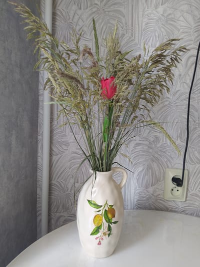 A bouquet of dried grass and a single pink flower stands in a cream-colored ceramic jug adorned with a painted lemon branch. The jug is placed on a white table. In the background, a wall with a tropical leaf pattern wallpaper is visible, along with a white pipe, a power outlet, and a black electrical cord. The scene is indoors, likely in a residential setting in Gusev, Russia. No people or animals are present. The lighting is natural, coming from an unseen source, casting soft shadows.