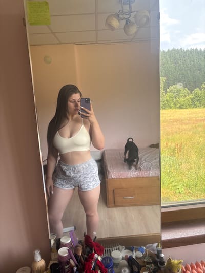 A young woman with long, dark hair is taking a mirror selfie in what appears to be a bedroom. She is wearing a cream-colored bralette and grey patterned shorts. She is holding her phone up to take the photo, and her nails are painted a light blue color. In the reflection of the mirror, a black dog is visible on a bed. The room has light pink walls, and a window shows a view of trees and a grassy field. There is also a dresser with several bottles and makeup brushes on it, and a light fixture with three lamps. The lighting suggests it is daytime.
