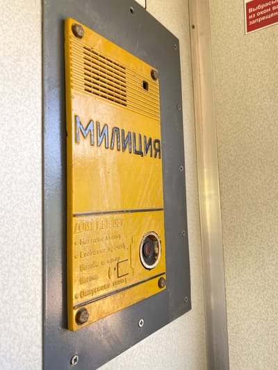 A close-up, eye-level shot shows a yellow intercom panel mounted on a gray metal plate attached to a textured, light-colored wall. The intercom panel is rectangular with rounded corners and features a speaker grill at the top. Below the grill, the word "МИЛИЦИЯ" (Militsiya, Russian for Police) is embossed in dark capital letters.

Beneath "МИЛИЦИЯ" are instructions in a smaller font. The first line reads "ДЛЯ ВЫЗОВА" (For Call), followed by a bulleted list:
* "Нажмите кнопку" (Press the button)
* "Сообщите причину," (Report the reason,)
* "Безбья и номер" (Your number)
* "Вагона" (Carriage)
* "Отпустите кнопку" (Release the button)

To the right of the instructions is a circular speaker or microphone grille with a dark center and a visible metallic edge. Below the speaker, a rectangular slot is depicted with an arrow pointing into it, suggesting a card or token insertion point.

The panel is secured to the gray plate with visible screws. The overall condition of the intercom appears aged, with some scuffs and wear visible on the yellow surface.

The environment surrounding the intercom is a narrow, interior space, possibly a public transport vehicle or building. A vertical metal trim piece is visible to the right of the intercom, separating it from the textured wall. The wall has a subtle, repeating pattern. A small red sign is partially visible in the upper right corner with white text, which appears to be a warning or informational notice. The specific location in the city of Kumuk, Russia is not discernible from the image itself.