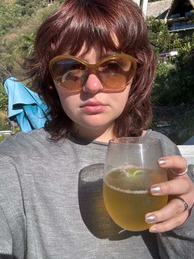 A person with reddish-brown, layered hair and bangs is depicted in a close-up, outdoors, under bright sunlight, within the city of San Pablo La Laguna, Guatemala. The individual wears large, amber-tinted cat-eye sunglasses and a small silver nose ring on the left nostril. They are dressed in a grey long-sleeved shirt. The right hand holds a clear, stemless glass containing a light amber carbonated beverage with ice and a lime wedge; the glass shows condensation. Fingernails on the right hand are painted metallic silver; a plain silver ring is on the ring finger, and a darker, textured ring with a green detail is on the pinky finger. In the background to the left, a light blue folded umbrella is visible, followed by a body of water and a steep, tree-covered hillside. To the right, behind the subject, a multi-level building with a distinctive thatched roof is visible on a lush green hillside.
