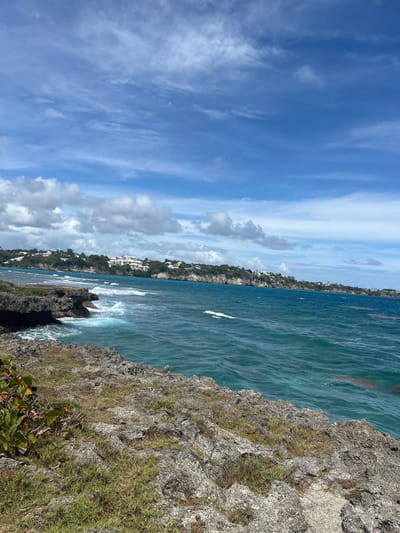 The image depicts a coastal scene near Le Gosier, France. The view is taken from a cliff overlooking the ocean. The water is turquoise-blue, with waves crashing against the rocky shoreline. In the distance, there is a coastline dotted with buildings, suggesting a residential or resort area. The sky is partly cloudy with streaks of blue visible. The cliff face is covered with vegetation, including grass and shrubs.