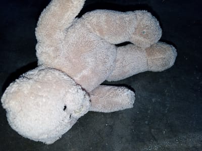 A light beige, plush teddy bear, appearing worn with flattened fur and some discoloration, lies horizontally on its back on a dark, textured surface. Its head faces the left, with two dark eye spots and a stitched facial feature visible. The bear's limbs are bent, with a small blue mark visible on its right upper leg. The dark surface, likely an unadorned floor, exhibits scattered dust, lint, and small particles, suggesting a utilitarian or aged environment. There are no other subjects, objects, actions, or interactions identifiable in the scene. The immediate environment is solely a dark, textured ground.