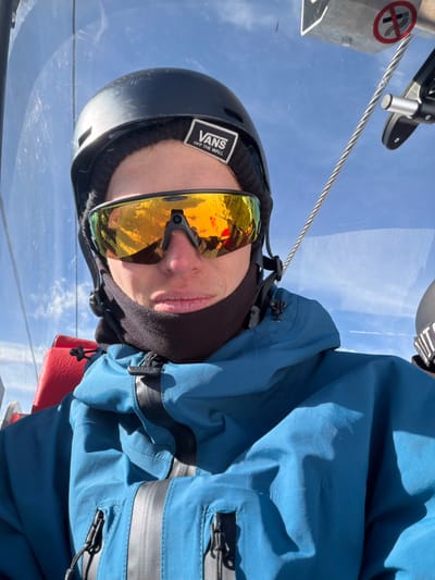 A sole individual is depicted in a close-up shot, seated within a ski lift cabin in Lunnaya Polyana, Russia. The person wears a black ski helmet, a black beanie with a white "VANS OFF THE WALL" patch centered on the forehead, and large reflective orange-gold sport sunglasses. A black fleece neck gaiter covers the lower face and neck. The individual is dressed in a teal blue waterproof ski jacket featuring multiple zippered pockets, some with black pull tabs. A red padded seat back is partially visible to the left of the person. Through the transparent cabin material on the right, metal components of the lift mechanism and a white sign with a red "no smoking" symbol are observable. The background consists of a clear blue sky with scattered white clouds. Reflections in the sunglasses show outdoor scenery.