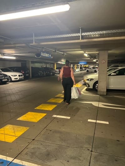 A person walks away from the camera in a dimly lit underground parking garage in Aachen, Germany. The person is wearing a dark red tank top and black pants, and is carrying a large white shopping bag. They are walking on a gray concrete floor with yellow and white painted lines and squares. Several cars are parked on either side of the aisle. Overhead, fluorescent lights illuminate the scene, and various pipes and conduits are visible along the ceiling. A blue sign with white text reading "Ausfahrt" and an arrow pointing to the right is mounted on the ceiling.