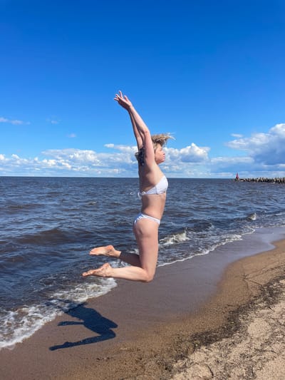 A full-body shot shows a woman in a white bikini jumping on a sandy beach next to the dark blue water of a large lake or sea. The sky is bright blue with scattered white clouds. The woman is in mid-air, with her arms raised above her head and her legs bent. Her body is in profile, facing to the right. The water has small waves crashing on the shore. In the distance, there is a breakwater made of large rocks and a red navigational marker. The sand is a light brown color and appears to be damp near the water's edge. The sun is casting a shadow of the woman on the sand. The scene is likely located in Belozersk, Russia, given the context provided by the prompt, which suggests it's a sunny day on the shore of a body of water.