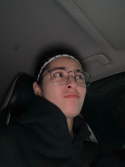 A single individual is depicted inside the interior of a vehicle in Komchén, Mexico, under low light conditions, possibly at night or during a rainy period. The person, appearing to be a young adult, occupies the left side of the frame, angled towards the right. They are wearing a dark hooded sweatshirt with a prominent collar, round metal-rimmed eyeglasses, and a white headband adorned with a repetitive dark pattern on their pulled-back dark hair. Their eyes are directed upwards and to the right, with a slight upward tilt of the head. The mouth is closed, with lips subtly pursed. A dark car seat headrest is visible behind and to the left of the individual. Above the person, the dark gray headliner of the vehicle is visible. To the right, a faint patterned reflection or condensation suggests a view through a window, possibly indicating rain outside. No other individuals or specific objects beyond those described are discernable.
