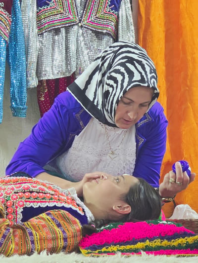 In Ribnovo, Bulgaria, two individuals are depicted in a close-up shot. The primary subject is a woman wearing a vibrant, traditional Bulgarian costume adorned with colorful embroidery and beads. Her head is tilted downwards, and her eyes are closed. She is lying on a textured surface, possibly a rug or blanket, which also features bright, woven patterns.
Positioned above her is another woman, whose face is partially obscured by a black and white zebra-print headscarf. She is wearing a royal blue jacket over a white lace top and a delicate gold necklace with a heart pendant. Her right hand is raised, and she is holding a small, dark blue object, possibly a cosmetic product or a decorative item. Her gaze is directed towards the woman below.
The background of the image includes glimpses of more traditional clothing hanging on the left, with sequins and intricate designs. An orange curtain is visible on the right, providing a warm contrast to the cooler tones of the clothing and the subjects. The overall impression is one of tradition, care, and possibly a ritual or grooming activity within a culturally rich setting.