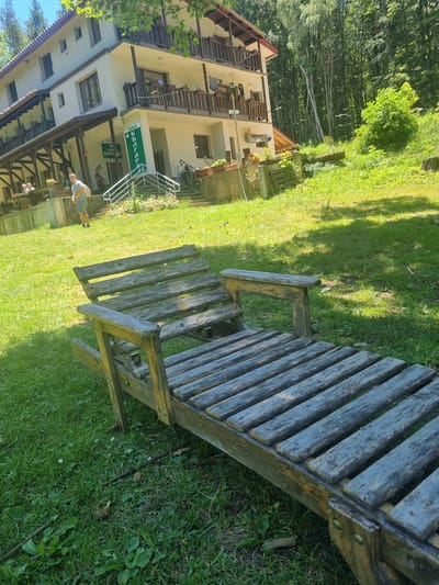 The image depicts a rustic wooden lounge chair in the foreground, set on a grassy lawn. Behind the chair, a three-story building with a white facade and wooden balconies is visible. A green sign with Cyrillic text, likely indicating the name of the establishment, hangs on the building. Several people are present on the building's grounds, some standing and others on the balconies. A bicycle is parked near the entrance. The environment is lush and green, with trees in the background suggesting a rural or mountainous setting, consistent with the location in Seltse, Bulgaria. The overall scene is one of a serene, outdoor setting at a guesthouse or similar accommodation.