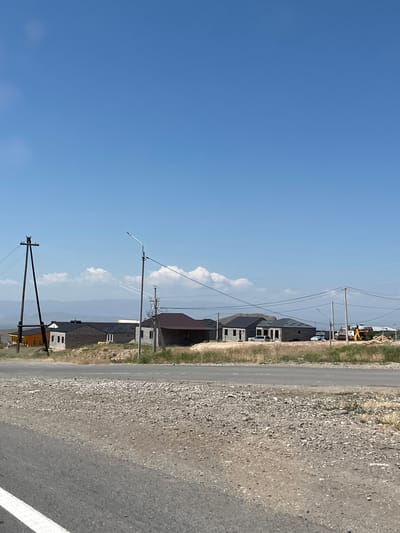 The image depicts a rural roadside scene in the vicinity of Zangakatun, Armenia, under a clear blue sky with scattered white clouds. In the foreground, a paved road with a white lane marker runs horizontally across the bottom. Adjacent to the road is a gravel shoulder.
The middle ground features a row of newly constructed or under-construction houses. Some houses are made of brick and stone, while others have dark-colored roofs. One house on the far left is painted orange. Several utility poles with wires are visible, including a prominent, dark wooden pole on the left. A street lamp is also present.
In the background, a distant mountainous landscape is visible, with a body of water or a valley separating the houses from the mountains. Construction activity is suggested by the presence of what appears to be an excavator or a similar piece of heavy machinery and piles of building materials on the right side of the image. The overall environment appears dry and arid. No people are clearly visible in the image.