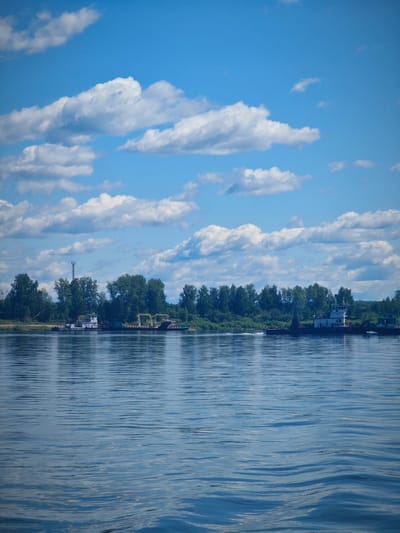 The image captures a wide, calm river on a bright, partly cloudy day. The water's surface is rippled, reflecting the sky and the lush green treeline on the opposite bank. Several boats are visible docked along the shore. One boat, a white tugboat, appears to be maneuvering, creating a wake. Another, larger vessel, possibly a barge or a ferry, is also present. A tall, lattice tower stands out against the trees on the left side of the frame. The sky is a vibrant blue, dotted with fluffy white clouds. The scene suggests a tranquil summer day at a riverside location, possibly near Krutoi Log, Russia, given the context.