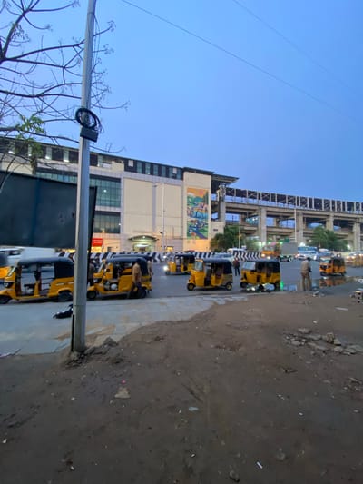 This image shows a street scene in St. Thomas Mount-Pallavaram Cantonment Board, India, likely during dusk or early evening due to the low light conditions and the blue hue of the sky. The foreground features a lamppost with some tangled wires.

The main subjects are a line of yellow auto-rickshaws parked along the side of a street. Several of these vehicles appear to be occupied by people, some of whom are visible through the windows. The rickshaws are lined up neatly, suggesting they are waiting for passengers.

In the background, a multi-story building with a modern facade is visible. It has large glass windows and appears to be a commercial establishment or an office building. Further down the street, another building with exposed concrete beams and balconies is under construction or renovation, adding a layered depth to the urban landscape.

The ground beside the parked rickshaws is a mixture of paved sidewalk and a rougher, dirtier area with scattered debris. The street itself appears to be somewhat wet, possibly from recent rain or cleaning.

There is a mural or large advertisement visible on the side of one of the buildings in the background, but the details are not clear enough to read any text. The overall atmosphere is that of a busy urban area at the end of the day, with the activity of transportation services still in motion.