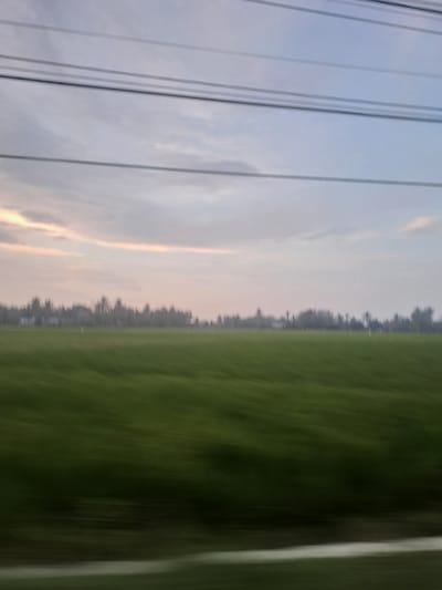 The image shows a blurred, sideways view. The left side features power lines against a light blue and pale orange sky, possibly at sunset or sunrise. The right side is dominated by green, blurred vegetation, likely a field or forest seen from a moving vehicle. A strip of what appears to be pavement is visible to the very right. The location is Lancoklancok, Indonesia, based on the prompt.
