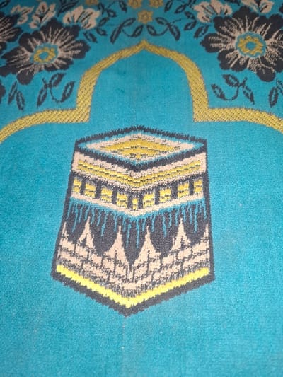 A turquoise-colored prayer mat is displayed in a close-up, overhead view, situated in Dan Maliki, Nigeria. The mat's central design features a stylized, multi-faceted representation of the Kaaba, rendered with geometric patterns in black, yellow, light blue, and beige tones. Above this central Kaaba motif, a prominent yellow arch pattern, indicative of a mihrab, is present. In the upper corners of the mat, flanking the mihrab, floral patterns with dark grey petals, yellow centers, and beige accents are visible against the turquoise fabric. The mat exhibits a woven textile texture. No individuals, actions, or direct interactions are visible within the frame.