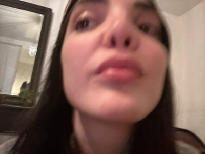 The scene, located in Roanne, France, depicts a close-up, blurry portrait of an individual's face, captured from a low perspective looking upwards. The subject, likely female, features dark hair, a prominent nose, and full lips. A light-colored headband is visible across the forehead, with dark hair framing the face. The eyes are partially visible near the top of the frame, directed slightly upwards. In the upper-left background, a dark-framed mirror reflects a white ceiling and a distinct white light fixture, which appears to have a feathery or spiky texture. Below the mirror, dark shapes and faint green foliage are discernible. The background to the right is a plain, light-colored wall. A patterned fabric, possibly part of the subject's clothing, is subtly visible at the bottom right.