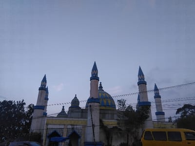 This image depicts a multi-domed building located in Kladno, Czechia, under an overcast, light grey-blue sky. The central structure features a large main dome and several smaller domes, all adorned with a distinct blue and yellow geometric pattern. The building's walls are primarily light-colored, possibly white or beige, with yellow accent details visible on its facade. Six slender, conical-topped towers, characteristic of minarets, rise from the building, painted in light beige with blue bases and blue and yellow patterned tops matching the domes. Dark green foliage from trees partially frames the building on both the left and right sides. Several dark utility lines traverse horizontally across the mid-ground, obscuring portions of the building and sky. In the lower right quadrant, the side profile of a bright yellow van with dark windows is partially visible, facing left. In the lower left quadrant, the dark roof of another vehicle is partially visible. No people are visible within the frame. The lighting is soft and diffused, indicative of an overcast day or twilight conditions.