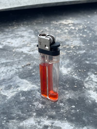 A clear plastic disposable lighter stands upright on a textured, dark gray surface, likely concrete or asphalt, at an unspecified location within Mns Asan, Indonesia. The lighter's transparent body reveals orange-red liquid fuel, which fills approximately half of its reservoir. The metallic components at the top, including the serrated flint wheel, show signs of wear and minor discoloration. A black plastic igniter lever is present. The supporting surface is uneven, covered with scattered light-colored granular debris, and exhibits subtle cracking. A blurred, lighter gray horizontal plane occupies the upper background. No individuals or active interactions are depicted.