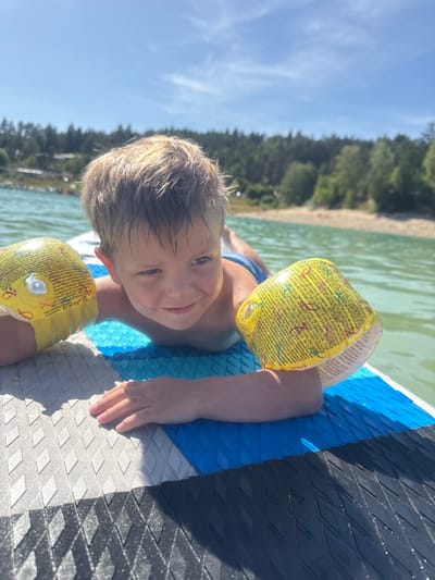 In Maisod, France, a young boy with blonde hair is pictured lying on a paddleboard in a lake. He is wearing yellow arm floaties and blue swim trunks. The water is a clear, greenish-blue, and a sandy shore with a dense forest line is visible in the background under a bright blue sky with scattered clouds. The paddleboard has a textured grip surface with blue, grey, and black sections. The boy is looking slightly off-camera with a relaxed expression.