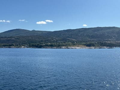 This media file depicts a scenic view of a body of water, likely a fjord or bay, in Bognes, Norway. The foreground is dominated by the expansive blue water, reflecting the sunlight with numerous small, sparkling glints. The water appears calm, with gentle ripples across its surface.
In the background, a verdant, tree-covered hillside rises steeply, forming a natural backdrop. Scattered amongst the trees and along the rocky shoreline are a few scattered houses, suggesting a sparse residential area. The hillside extends to meet a range of rolling mountains in the distance. The highest peak in the left portion of the image features a prominent tower, possibly a telecommunications mast. The sky is a clear, bright blue, with a few small, wispy white clouds scattered across it.
The scene is bathed in bright sunlight, indicating it is daytime, likely midday or early afternoon, given the angle of the light and the clarity of the sky. The weather appears to be pleasant and sunny, typical of a summer day in this region. The overall impression is one of natural beauty and tranquility. There is no visible text within the image.
