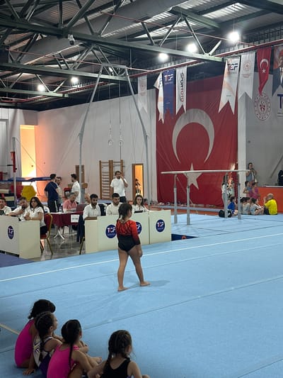 The image depicts a gymnastics competition at the Sancaktepe Municipality Sports Complex in Sancaktepe, Turkey. In the foreground, several young girls in leotards are seated on a blue mat, watching the competition. A female gymnast in a black and red leotard stands on the mat, facing away from the camera. In the midground, a set of parallel bars is visible. Officials and spectators are seated at tables behind the bars, some wearing white shirts. A large Turkish flag hangs in the background, along with banners displaying the TCF (Turkish Gymnastics Federation) logo. The environment appears to be an indoor sports facility with a high ceiling, exposed metal beams, and bright lighting.