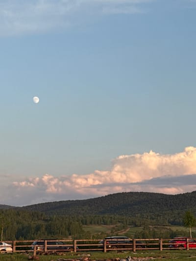 The image shows a landscape in Кузнецово, Russia. The sky is predominantly blue with clouds on the right side of the image. The moon is visible in the lower left quadrant. Adjacent to the clouds is a densely forested hill. On the right, a fence is visible, with cars parked along it.