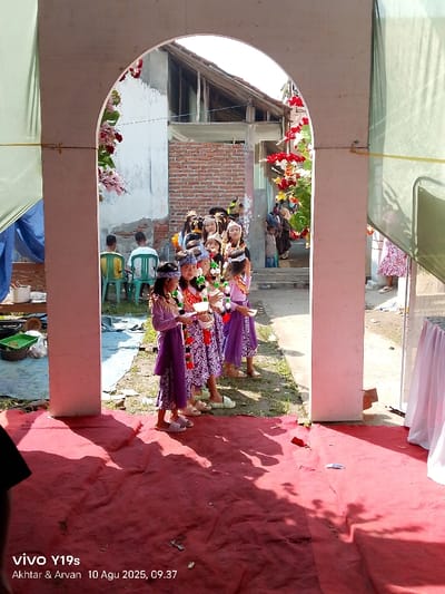An outdoor scene in Jagapura Kulon, Indonesia, is framed by a light-colored arched entryway. The foreground is covered by a red carpet. Through the archway, approximately twelve young girls are positioned in a line, facing the viewer. The four closest girls wear matching purple long-sleeved tops, patterned sarong-like lower garments, headbands, and garlands. They each hold a small white bowl or cup in their hands.
Behind the girls, various individuals are visible. Two men sit on green plastic chairs on the left side of the background. Further back on the right, additional people, including a child in a blue shirt, are present. The environment beyond the archway features unpainted concrete walls, a partially visible brick wall, and building structures with visible roof framing. The archway itself is adorned with red and white artificial floral decorations on the right side and pink ones on the left. The ground outside the arch appears to be unpaved dirt. The scene is brightly lit by natural sunlight, casting shadows across the red carpet in the foreground.