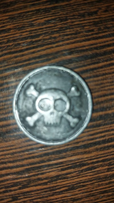 A circular, metallic object with a skull and crossbones motif is centered on a wooden surface. The wood grain displays horizontal lines of alternating brown and black. The metallic object is silver-toned and appears to be a coin or a medallion, with the raised design of a skull in the center, flanked by two crossed bones. The lighting in the image creates a soft glare on the surface of the object. No specific location in Kansk, Russia can be determined from the provided image.