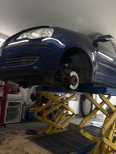 A blue car is elevated on a yellow hydraulic lift in what appears to be an automotive repair shop in Rakovica Urban Municipality, Serbia. The front driver-side wheel has been removed, exposing the brake rotor and suspension components. The car's headlight and grille are visible. Various tools and equipment are visible in the background, along with a white tiled wall.