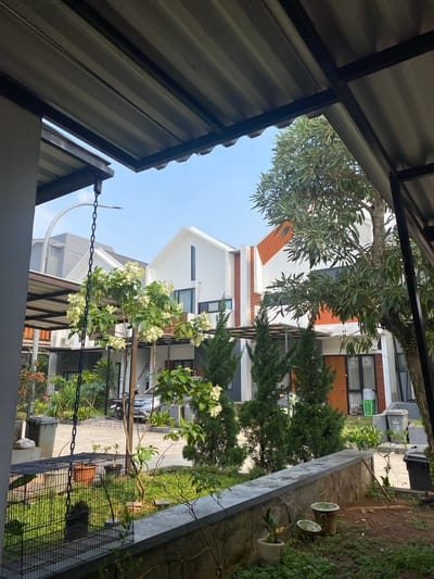The image shows a residential area in Cibinong, Indonesia. In the foreground, a metal cage is partially visible, with lush green grass and potted plants around it. A concrete ledge runs along the bottom of the frame. Behind this, a row of modern houses with white and orange facades are visible. Trees and lush greenery are scattered throughout the scene, including a flowering bush with white blossoms and a large tree with dense foliage. A silver car is parked on the paved driveway. The sky is clear and blue. The structures are typical of suburban housing in Indonesia, characterized by their modern architecture and well-maintained landscaping.