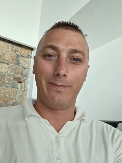 A medium shot captures a middle-aged Caucasian man with fair skin and a short, spiky haircut. He has light blue eyes and a slightly pursed lip expression. He is wearing a white collared shirt with visible buttons. A thin silver chain is around his neck. The background is split between a textured stone wall on the left and a plain white wall on the right. The photo was taken indoors. The subject is in the Gouves Municipal Unit, Greece, as indicated by the broader context, though specific location details within the municipality are not evident from the image.