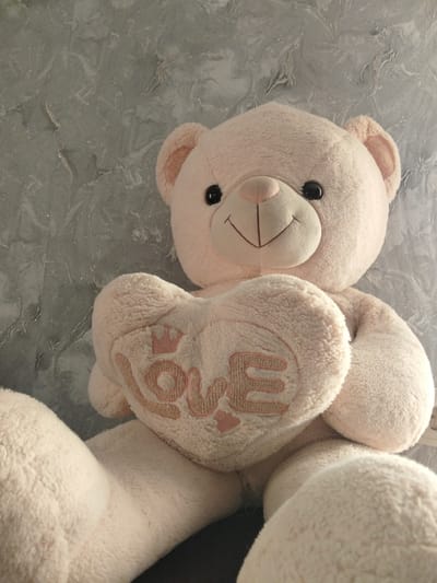 This media file features a close-up, eye-level shot of a light pink teddy bear. The bear is positioned in the center of the frame, with its head tilted slightly to the left. It is holding a large, light pink heart-shaped pillow with the word "love" embroidered in a darker pink thread, along with a small crown above the "o". The bear has black button eyes, a stitched nose, and a stitched smiling mouth. Its ears are slightly rounded and positioned on the sides of its head.
The background is a gray, textured surface with a marbled pattern, appearing to be a wall or a decorative panel. The lighting is soft and diffused, suggesting an indoor setting, possibly during the day. The overall mood is gentle and affectionate, reinforced by the teddy bear and the heart-shaped pillow. There is no visible information about the time of day, weather, or specific location beyond the general context of Birsk, Russia. The primary subjects are the teddy bear and the heart-shaped pillow. There is no discernible activity or event taking place, as the image is a still portrait of the object.