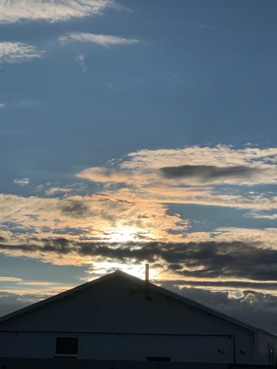 The image captures a sunset over a building in Большое Афанасово, Russia. The sky is filled with dramatic clouds, with the sun peeking through, casting golden and orange hues. The silhouette of a house, with its pitched roof and a chimney, dominates the foreground. The building appears to be a simple structure, possibly a residential home or a barn. The lighting suggests it is late afternoon or early evening. There are no people visible in the image, and the environment appears to be rural or suburban.