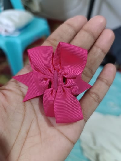 A close-up shot shows a person's hand holding a bright pink ribbon bow. The bow is elaborately crafted with multiple loops and pointed ends, giving it a star-like appearance. The hand is open, with the palm and fingers visible, providing a textured background for the accessory. The lighting is soft, highlighting the sheen of the ribbon and the lines on the hand. In the blurred background, a turquoise object and white fabric are faintly visible, suggesting an indoor setting. The specific location in Kawit, Philippines is not identifiable from the visual information provided.