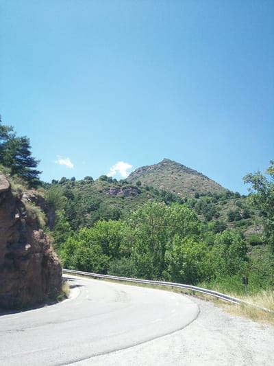 The image depicts a winding road curving through a mountainous, forested landscape under a clear blue sky. The road is paved and has white lane markings. A metal guardrail runs along the edge of the road, separating it from the steep, rocky incline on the left and the dense vegetation on the right. The vegetation consists of a variety of trees and shrubs, with a prominent peak of a mountain dominating the background. There are a few scattered white clouds in the sky. The scene appears to be located in a natural, undeveloped area, likely a mountain pass or a rural route. There are no people or animals visible in the image. The lighting suggests it is daytime, possibly mid-morning or afternoon, given the bright sunlight. The location is identified as being in the city of Berga, Spain, which is known for its mountainous terrain and natural beauty.