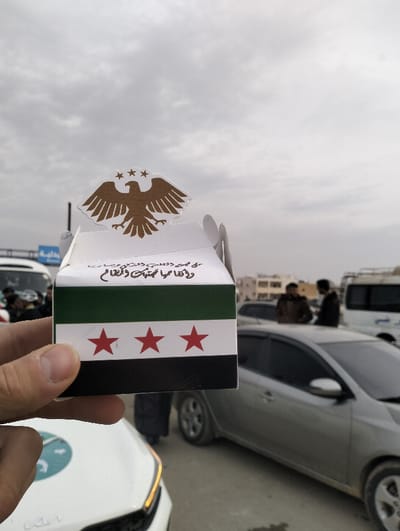 A hand holds a white box decorated with the flag of Syria, featuring a green, white, and black horizontal tricolor with three red stars. A golden eagle with three stars above its head adorns the top. Arabic script is visible below the eagle. The background shows an outdoor urban scene with cars, people, and cloudy skies. A white Kia is partially visible in the foreground. Two men stand near a parked silver car. Other vehicles, including a white van and a bus, are further back.