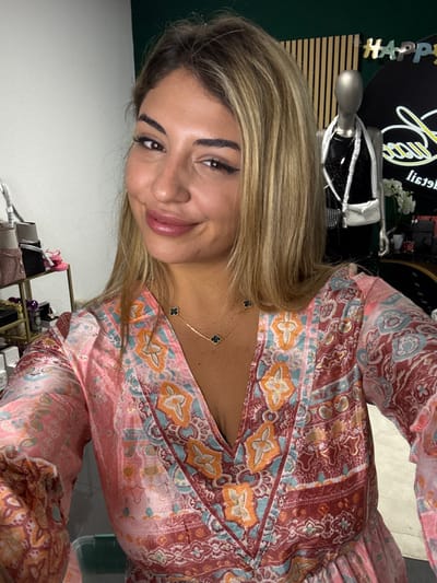 A young woman with blonde hair, styled with dark eyeliner, poses in a patterned dress. Her neckline reveals a delicate gold chain necklace adorned with black clover-shaped pendants. The dress features a mix of pink, orange, and turquoise patterns. Behind her, a mannequin torso, and decorative objects are visible against a dark background with gold accents. The backdrop appears to be a retail setting, possibly a clothing store.