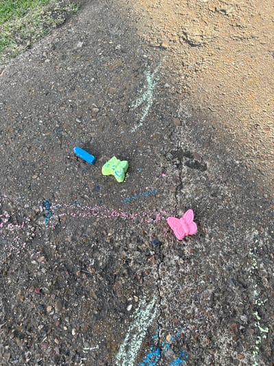 The image depicts a section of asphalt pavement with scattered chalk drawings and chalk pieces. A single blue chalk stick lies on the ground. A green chalk drawing in the shape of a butterfly is present, accompanied by some pink and blue chalk dust. Further along the pavement, a pink chalk butterfly is visible. Streaks of white, blue, and pink chalk mark the asphalt in various places. The pavement shows cracks and is a mix of dark gray and lighter brown tones. To the left of the pavement, a strip of green grass is visible, indicating an outdoor urban environment. The overall scene suggests recent children's activity in a public space.
