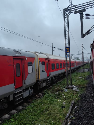 The image shows a red and grey train parked on a track next to a grassy embankment littered with debris. The sky is overcast and grey. A tall metal pole with overhead electrical wires stands to the right of the train. A blue sign with a yellow "M" symbol and text in Hindi is attached to the pole. The train appears to be at a station, as suggested by the platform and signage. The overall scene depicts a typical railway environment in India, likely on a cloudy day. Based on the distinct signage and train livery, this scene is identifiable as being in Bhopal, India.