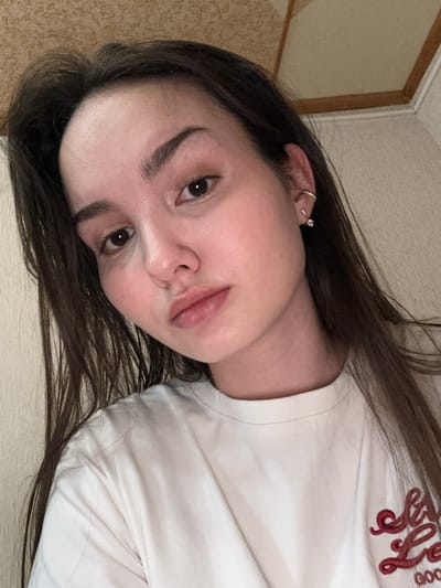 A single female individual is depicted in a head-and-shoulder portrait indoors. The subject has fair skin, dark brown hair, and dark brown eyes, looking directly into the camera. Her lips are slightly parted in a pout. She wears a small hoop earring in the helix of her visible left ear and a stud earring with a clear stone in her earlobe. Her attire consists of a light-colored, possibly white or off-white, crew-neck t-shirt. Partial red embroidery is visible on the right side of the t-shirt, appearing to spell "Stay Good" or similar text. The background features a light-textured wall and a ceiling with a light-colored panel and dark wooden trim. The specific location within Bayronovka, Russia, cannot be determined from the visual information provided. The image is a self-portrait.