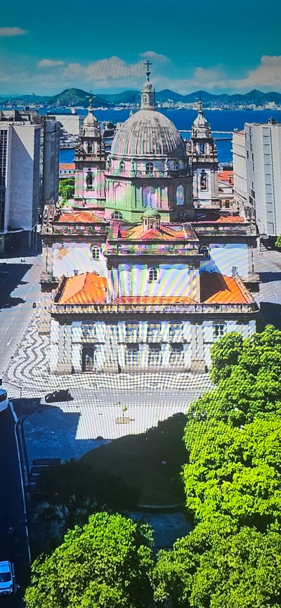 This is a high-angle, aerial view of a large, ornate domed building with a green dome, flanked by smaller towers and multiple levels of architecture. The building is constructed of stone and features numerous windows and balconies. It is situated in a city environment, surrounded by modern, tall buildings. In the foreground and to the right, a dense canopy of lush green trees dominates the lower portion of the image. The background reveals a body of water, likely a bay or harbor, with distant hills and mountains under a bright, partially cloudy blue sky. The street in front of the building has a patterned paving design and a few vehicles, including a dark-colored motorcycle and a white van. The overall impression is one of a historic architectural landmark within a bustling urban landscape, adjacent to a natural waterfront and abundant greenery. The image has some digital artifacts, appearing as vertical lines and color distortions, particularly over the building and sky.

The location of Lippstadt, Germany is not visible in this image.