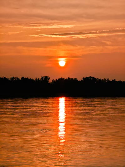 This is a vertical, medium-shot photograph of a sunset over a body of water in Maloe Nakhval'skoe, Russia. The sun is a bright orb, slightly off-center to the left, casting a warm, orange glow across the sky and reflecting on the water. The sky is filled with wispy clouds, also tinged with orange and red hues. A dark silhouette of a tree line forms the horizon between the sky and the water. The water itself is calm, with subtle ripples that catch the sunlight, creating a shimmering path from the sun to the foreground. No people or animals are visible in the frame.