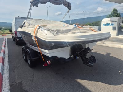 A white and dark gray boat is on a trailer, parked on an asphalt surface. The boat is covered with a light gray tarp secured by orange straps. A white van is parked to the left of the boat. In the background, there is a Lukoil AdBlue dispenser and a few trees, with mountains visible in the distance under a cloudy sky. The scene appears to be a gas station in Chuchuligovo, Bulgaria.