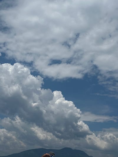 The image shows a daytime view of the sky with a mix of cumulus and stratus clouds. Patches of blue sky are visible between the clouds. A dark mountain silhouette is on the right. The location is Ortaca, Turkey.